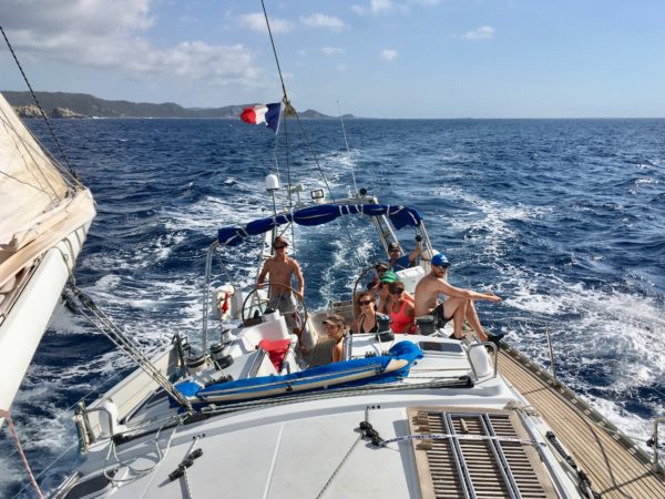 Un équipage heureux dans le cokpit d'un voilier, sous le soleil et sous une bonne brise, avec la côte de la Corse occidentale en arrière plan.